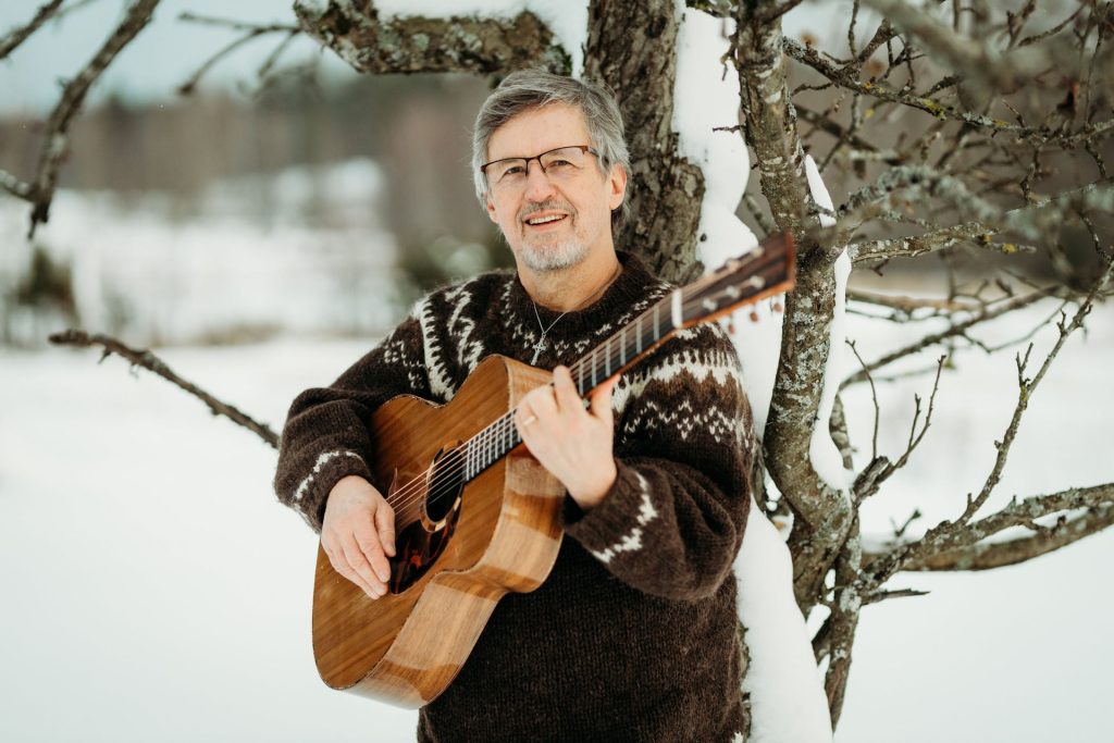 Kuvassa Pekka Simojoki kitara kädessä puun edessä.