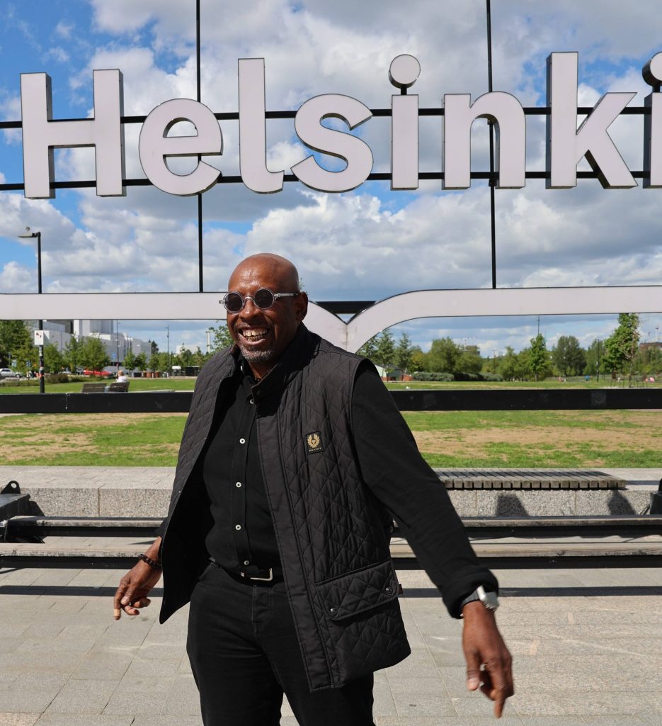 Kienda Hoji seisomassa Helsinki-kyltin edessä aurinkoisessa säässä.